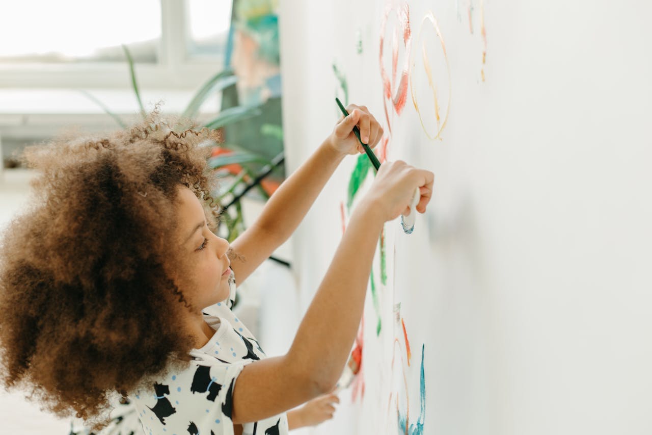 hero-services A young girl with curly hair paints creatively on a white wall in a bright indoor setting.