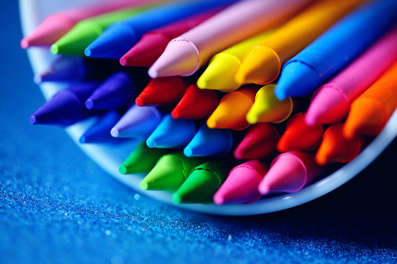 impact-img A close-up of vibrant colored crayons arranged in a bowl on a blue surface.